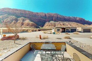Een terras met een tafel en stoelen, een gebouw met een garage, en een bergachtige achtergrond.