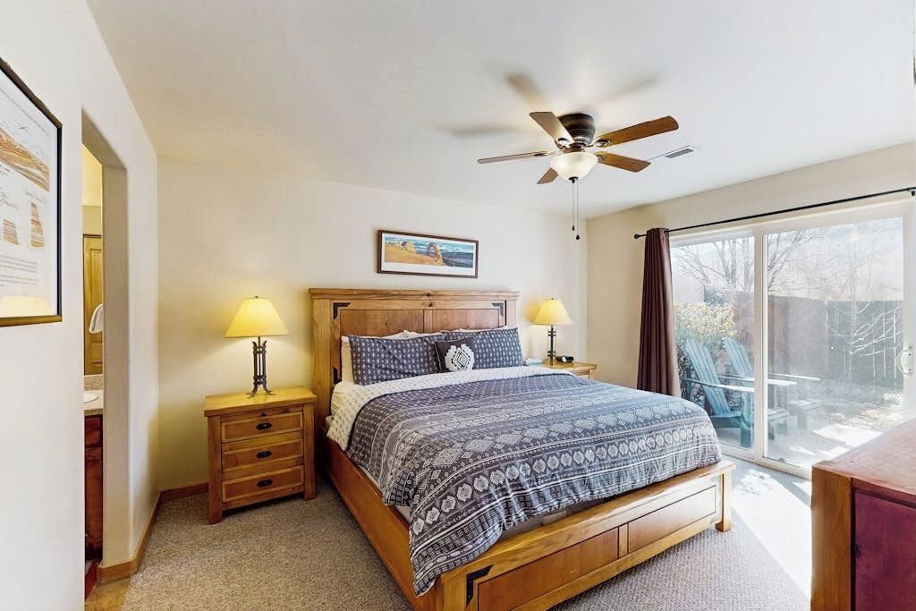 A bedroom with a bed, two bedside tables with lamps, a ceiling fan, a window with curtains, and a framed picture on the wall.
