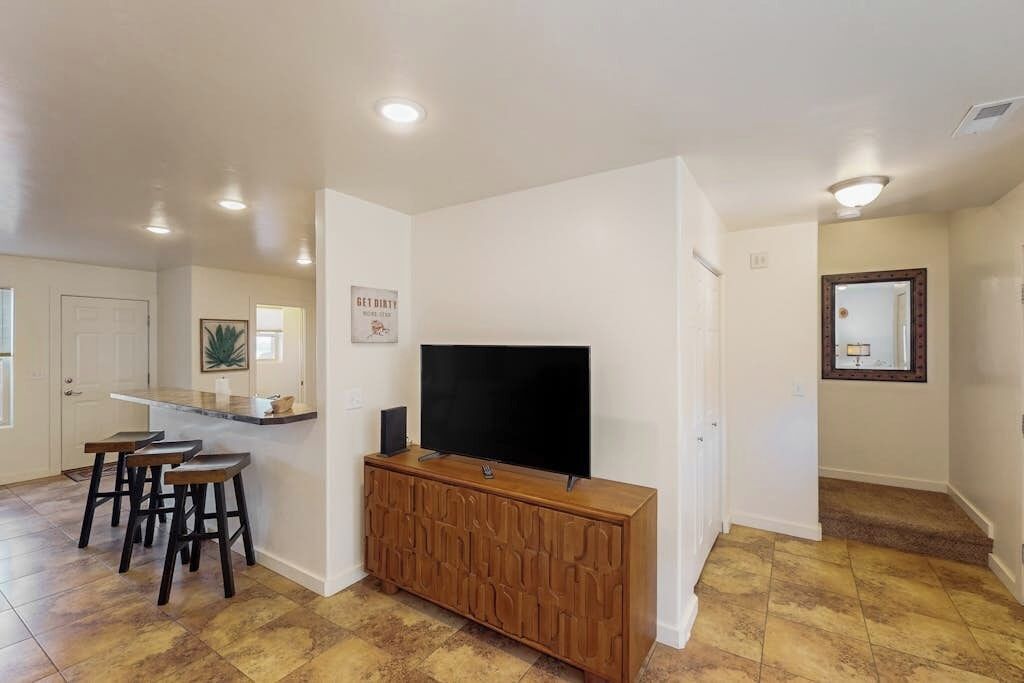 Una sala de estar con un televisor de pantalla plana sobre un mueble de madera, un área de cocina con un taburete de bar y un pasillo con un espejo.