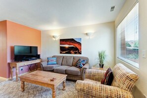 Ein Wohnzimmer mit Flachbildfernseher, einer Couch, einem gemusterten Sessel, einem Couchtisch und einem Bild an der Wand.