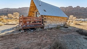 Una cabaña de madera con techo metálico, rodeada por un paisaje árido con montañas al fondo.