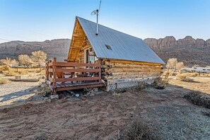 Una cabaña de madera con techo metálico, rodeada por un paisaje árido con montañas al fondo.