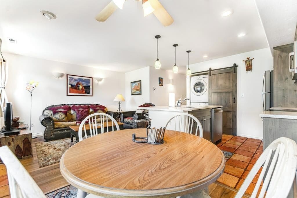 A dining area with a round wooden table and white chairs, a sofa with a patterned throw, a washing machine, and a kitchen area with a sink and cabinets.