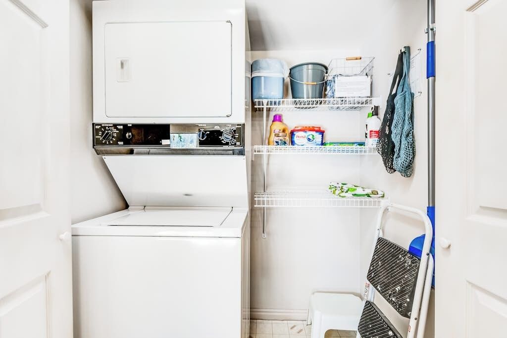 Une petite buanderie comprenant un lave-linge, des étagères avec des produits de nettoyage et un étendoir.