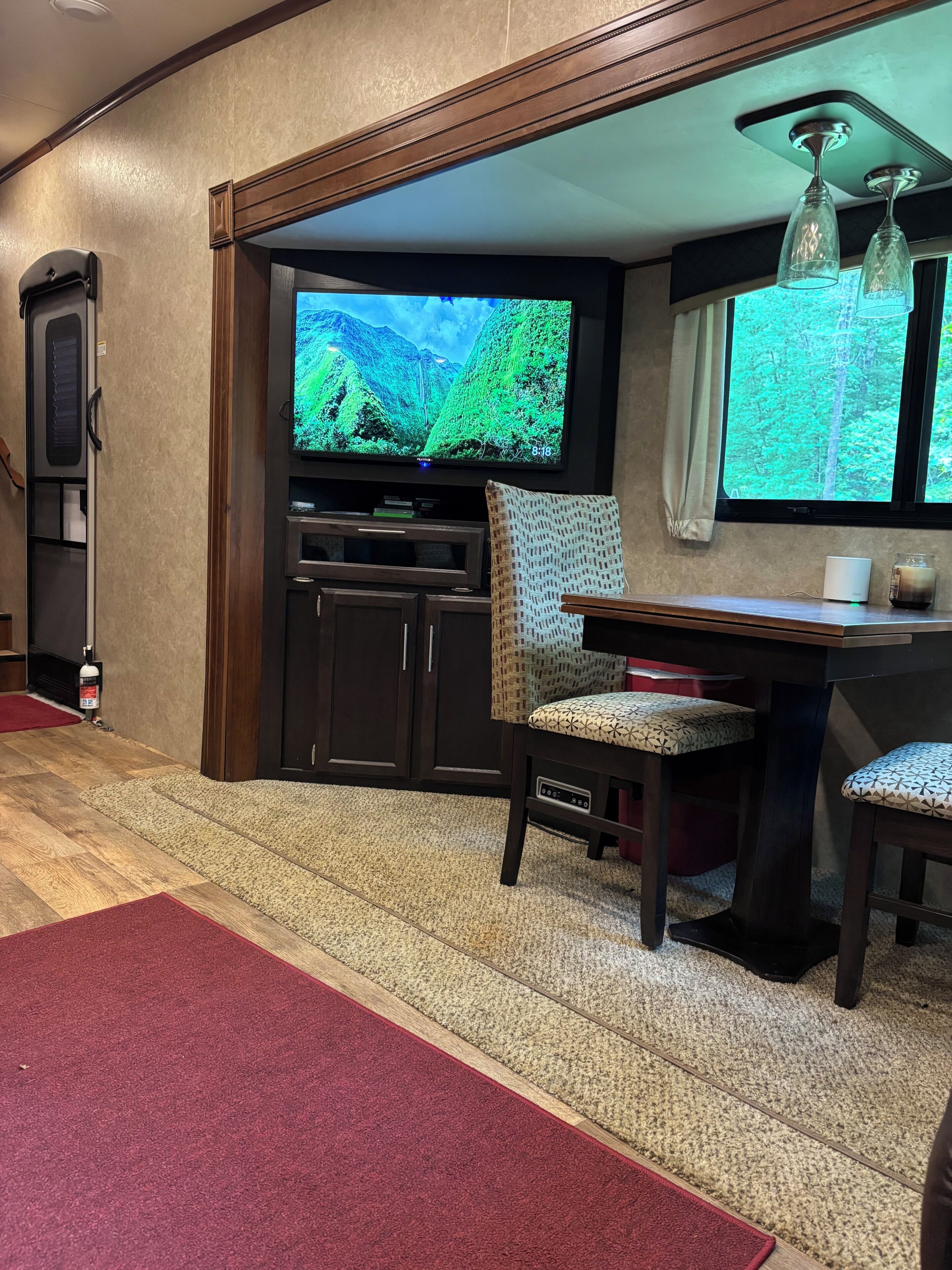 Ein Zimmer mit Fernseher, der eine Berglandschaft zeigt, einer Sitzecke mit Tisch und Stühlen und einem roten Teppich, der zu einer Tür führt.