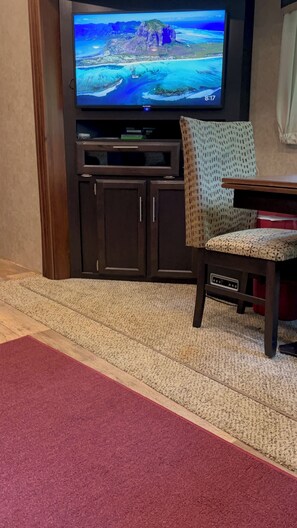 A living room with a flat-screen TV mounted on a dark wooden cabinet, a patterned rug, and a red carpet leading to a dining area.