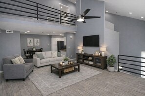 A spacious living room with a high ceiling, a ceiling fan, a flat-screen TV, a sofa, a coffee table, and a dining area with a table and chairs. - Sun Devil at 5th & Mill - 3BR - ASU, Mill Ave & Spring Training (Tempe)
