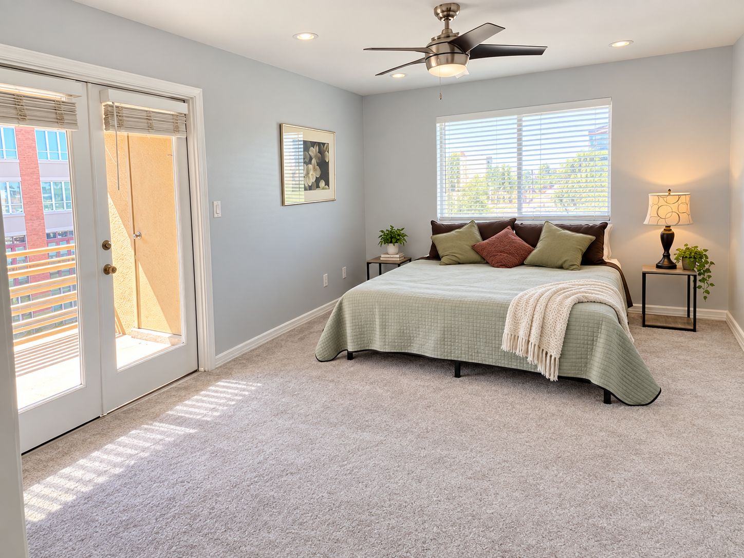 A bedroom with a bed, a ceiling fan, a window with blinds, a door, and a small table with a lamp.