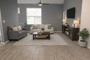 Ein Wohnzimmer mit grauem Sofa, beigem Sofa, Couchtisch aus Holz, Wandfernseher und einer Topfpflanze.