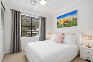 A bedroom with a bed, curtains, a window, a nightstand, a lamp, and a painting on the wall. - Spacious 3-Bedroom Townhouse Near Terrigal Beach (Terrigal)