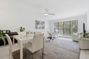 Ein modernes Wohnzimmer mit einem Glastisch, weißen Sofas und einem großen Fenster mit Jalousien.