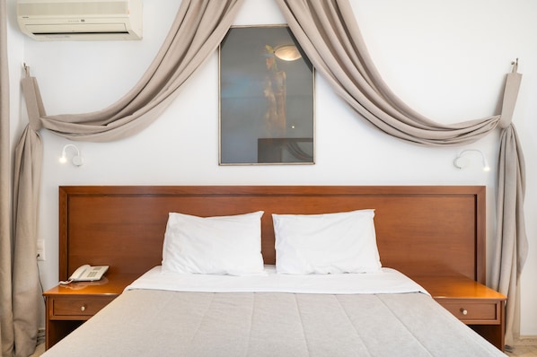 Habitación de hotel con cabecera de madera, cama con sábanas blancas, teléfono junto a la cama y aire acondicionado instalado en la pared.