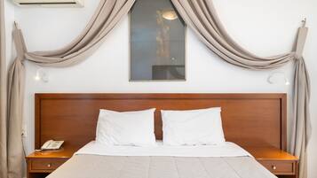 Quarto de hotel com cabeceira de madeira, cama com roupa de cama branca, telefone na mesinha de cabeceira e ar condicionado fixado na parede.
