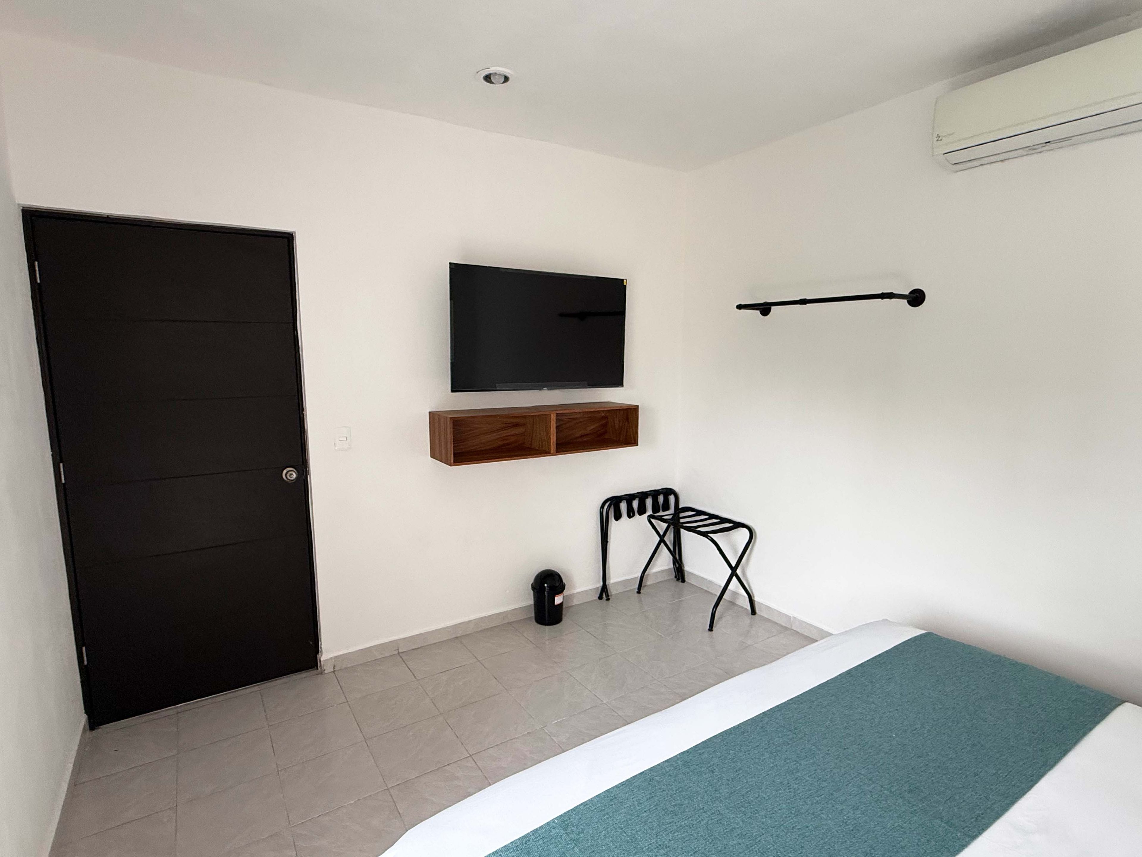 Une chambre à coucher avec un lit, une porte noire, un téléviseur fixé au mur, une petite étagère en bois, une chaise noire et un appareil de climatisation.