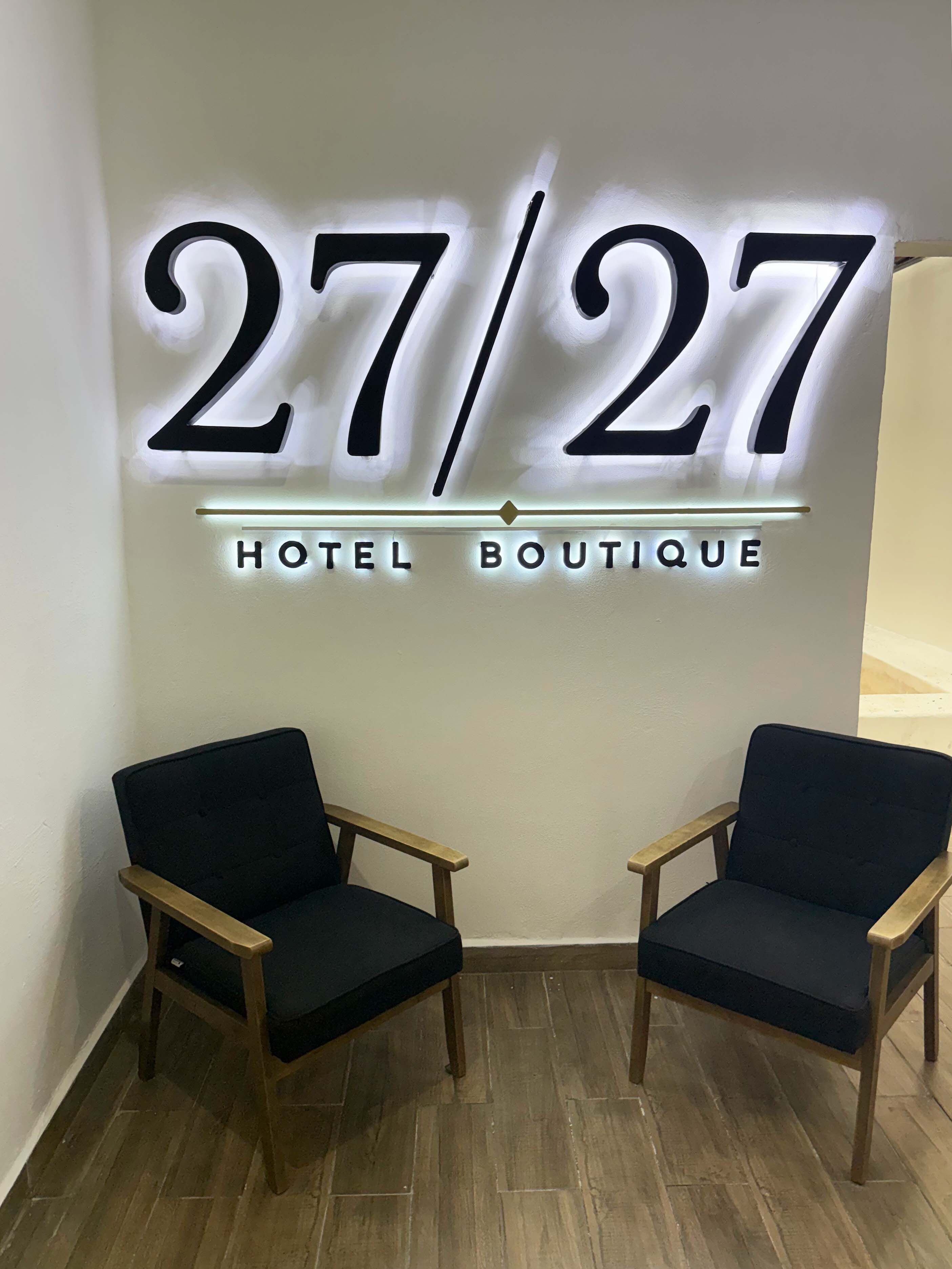 Ein Hotel-Schild mit den Zahlen '27/27' und dem Schriftzug 'HOTEL BOUTIQUE' in beleuchteten Buchstaben. Vor dem Schild stehen zwei Holzstühle mit schwarzen Kissen.