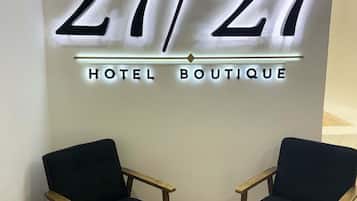 A hotel sign with the numbers '27/27' and the words 'HOTEL BOUTIQUE' in illuminated letters. There are two wooden chairs with black cushions in front of the sign.