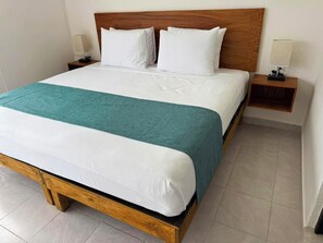 Uma cama com estrutura de madeira, roupa de cama branca e um cobertor azul-turquesa. Há duas mesas de cabeceira com abajures.