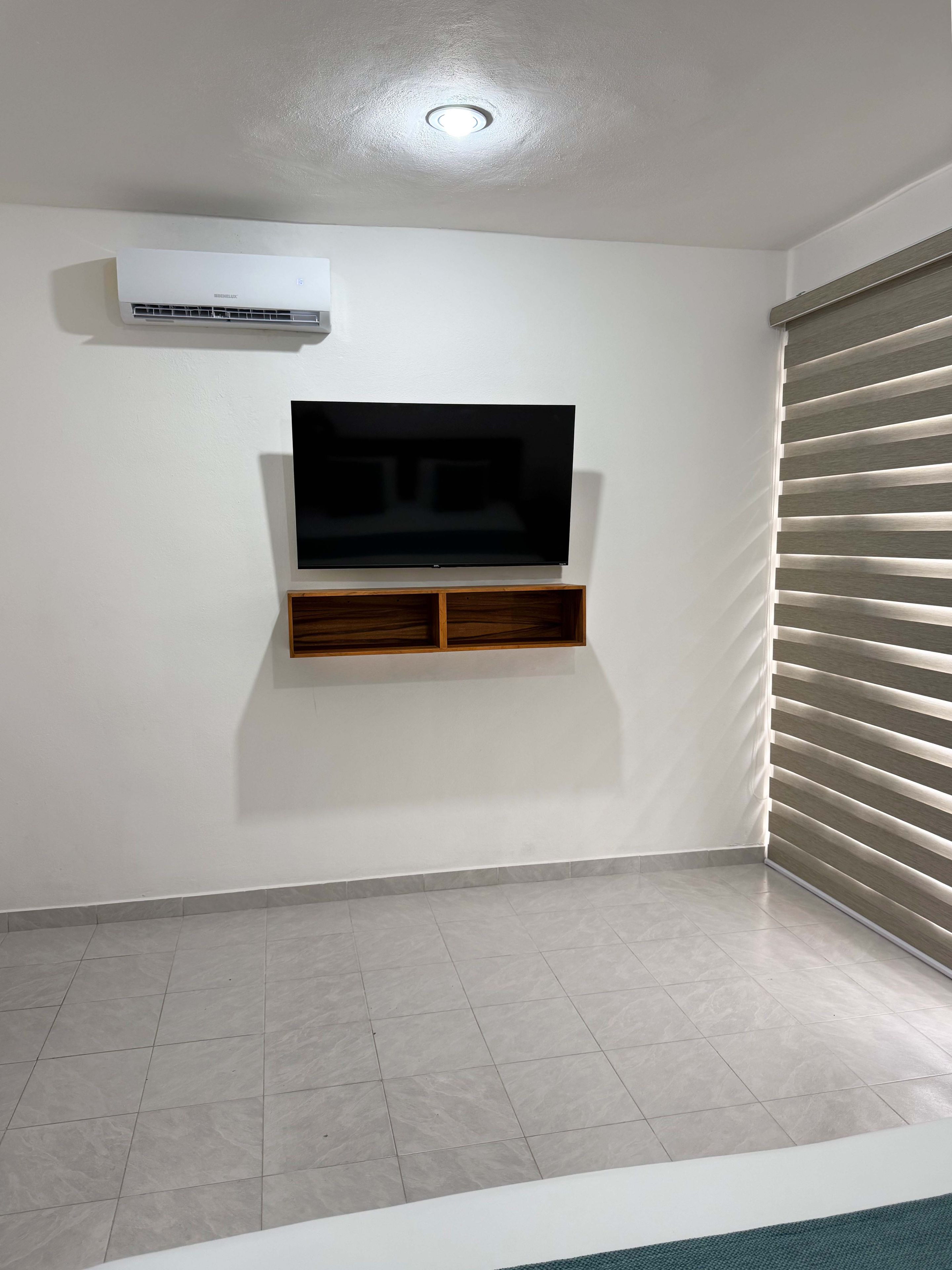 Une pièce dotée d’un téléviseur à écran plat fixé au mur, d’un appareil de climatisation placé au-dessus, et d’une fenêtre avec des stores.