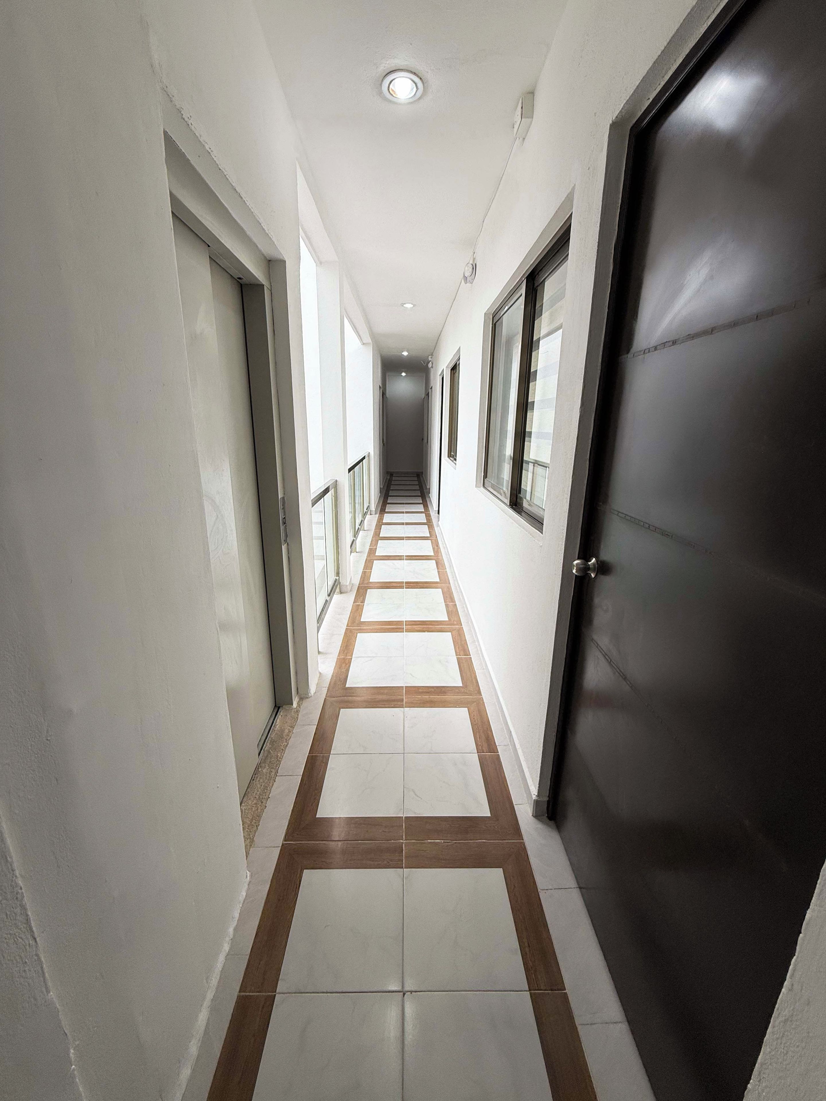 Un couloir au sol carrelé, avec une porte sur la droite et des fenêtres sur la gauche.