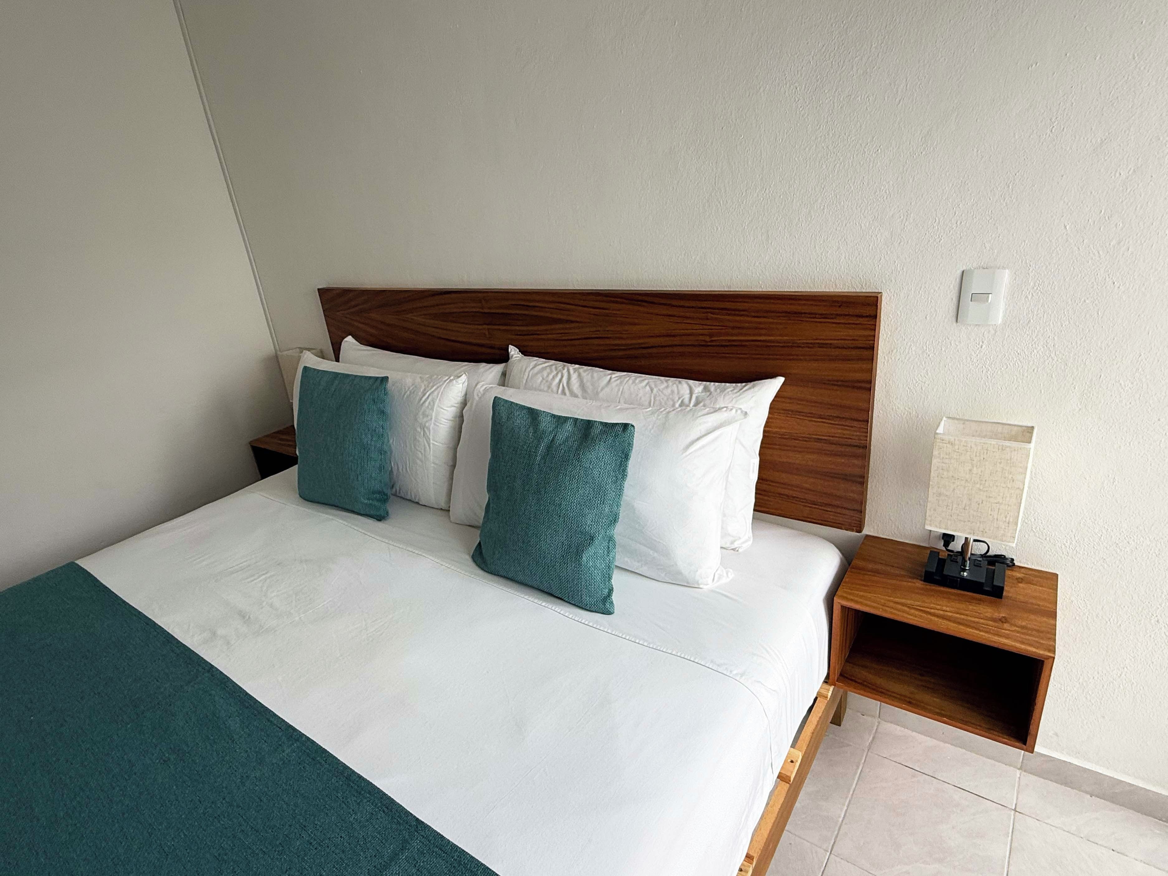 Ein ordentlich bezogenes Bett mit weißen und türkisfarbenen Kissen, ein hölzernes Kopfteil, ein Nachttisch mit Lampe und ein Lichtschalter an der Wand.
