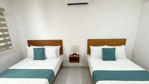 Een hotelkamer met twee eenpersoonsbedden, elk met een houten hoofdbord, wit beddengoed en turquoise kussens. Daartussen staat een klein nachtkastje met een lamp.