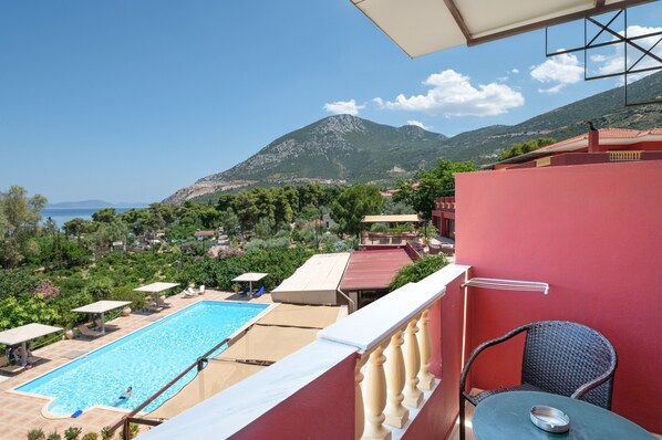 Vista de la piscina con montañas al fondo, zona de comedor y un edificio rojo.