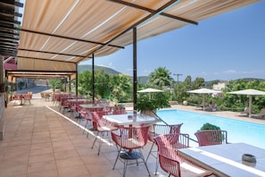 Un espacio al aire libre techado con mesas y sillas, piscina y vistas a las colinas.