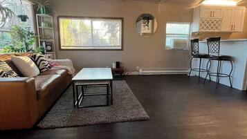 A living room with a sofa, a coffee table, and a dining area with chairs. There are windows with blinds, a mirror, and a kitchen area with cabinets.