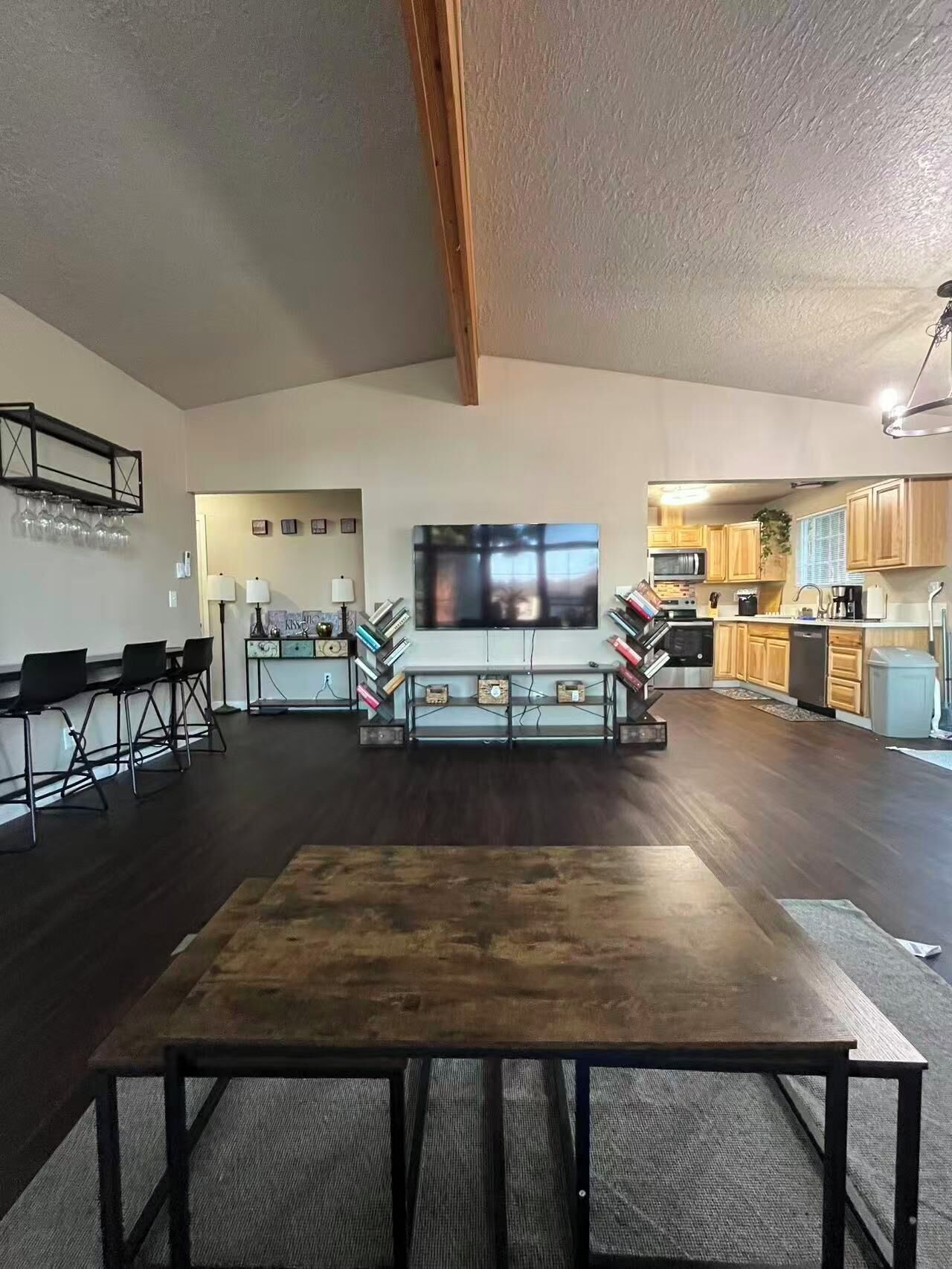 A spacious living area with a large flat-screen TV, a wooden dining table, and a kitchen in the background.