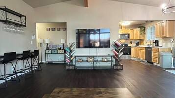 A spacious living area with a large flat-screen TV, a wooden dining table, and a kitchen in the background.