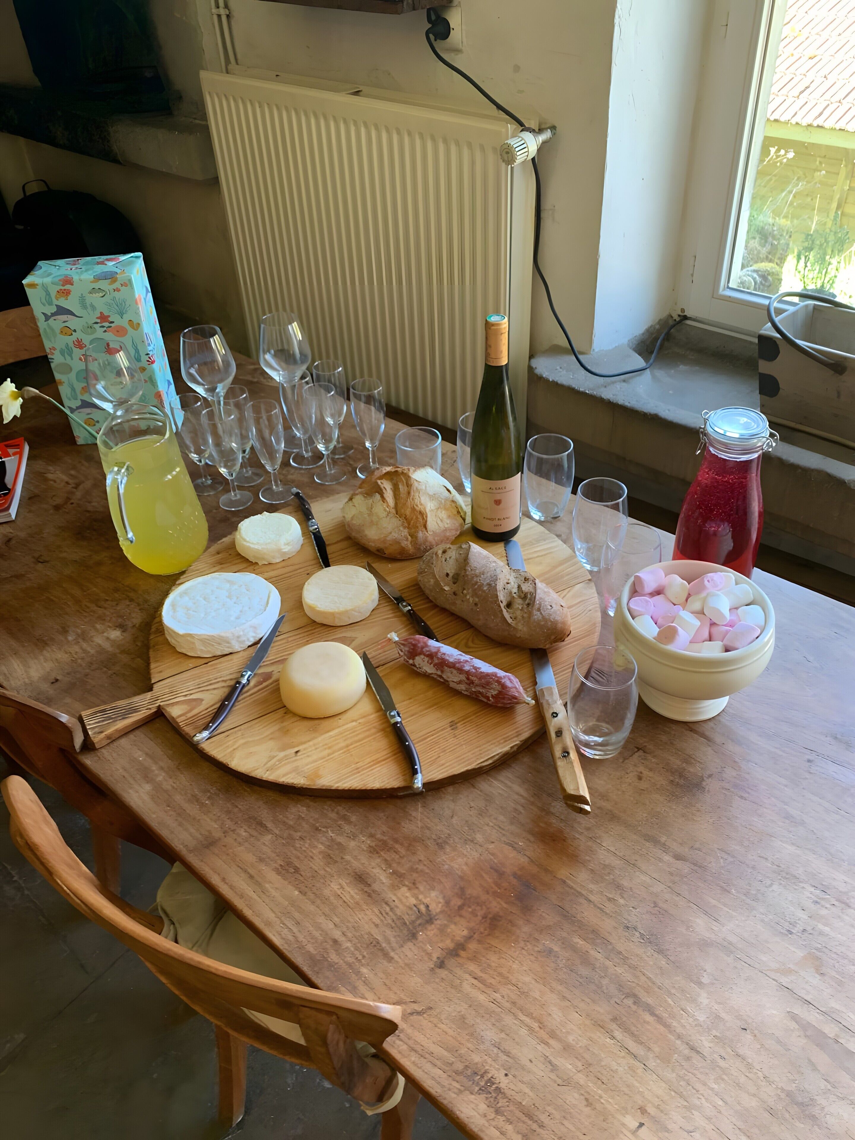Une table en bois sur laquelle sont disposés divers aliments, notamment du fromage, du pain et une bouteille de vin, ainsi que plusieurs verres et une carafe contenant une boisson jaune.