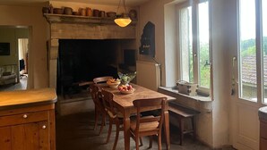 A kitchen with a fireplace, wooden dining table, chairs, and a window with a view. - Holiday Home "Harmangouty": Private Pool, 2.5 ha Grounds & Tranquil Forest Retreat (Raon-aux-Bois)