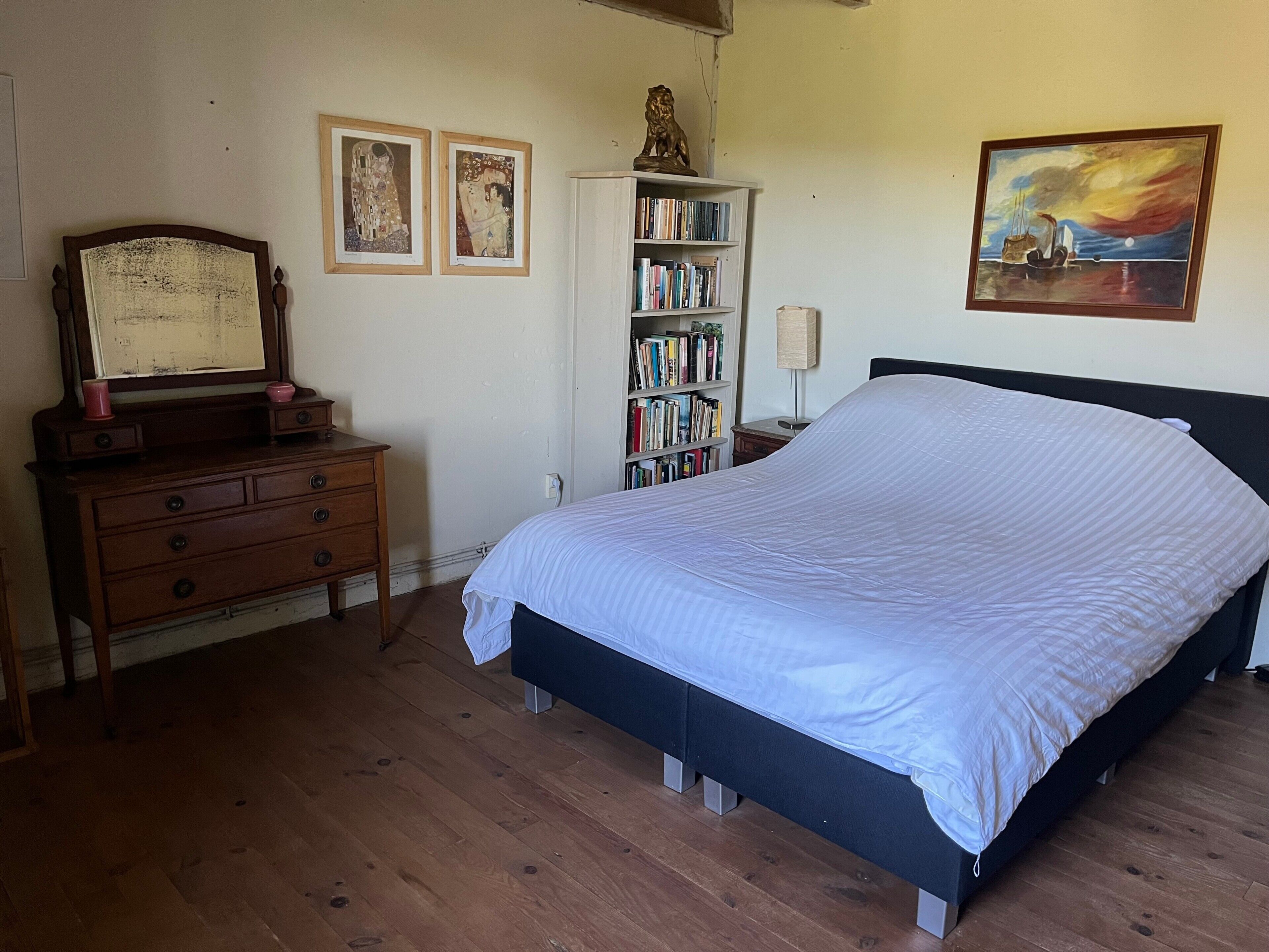 Une chambre à coucher comprenant un lit, une commode, une bibliothèque et deux œuvres d’art encadrées accrochées au mur.