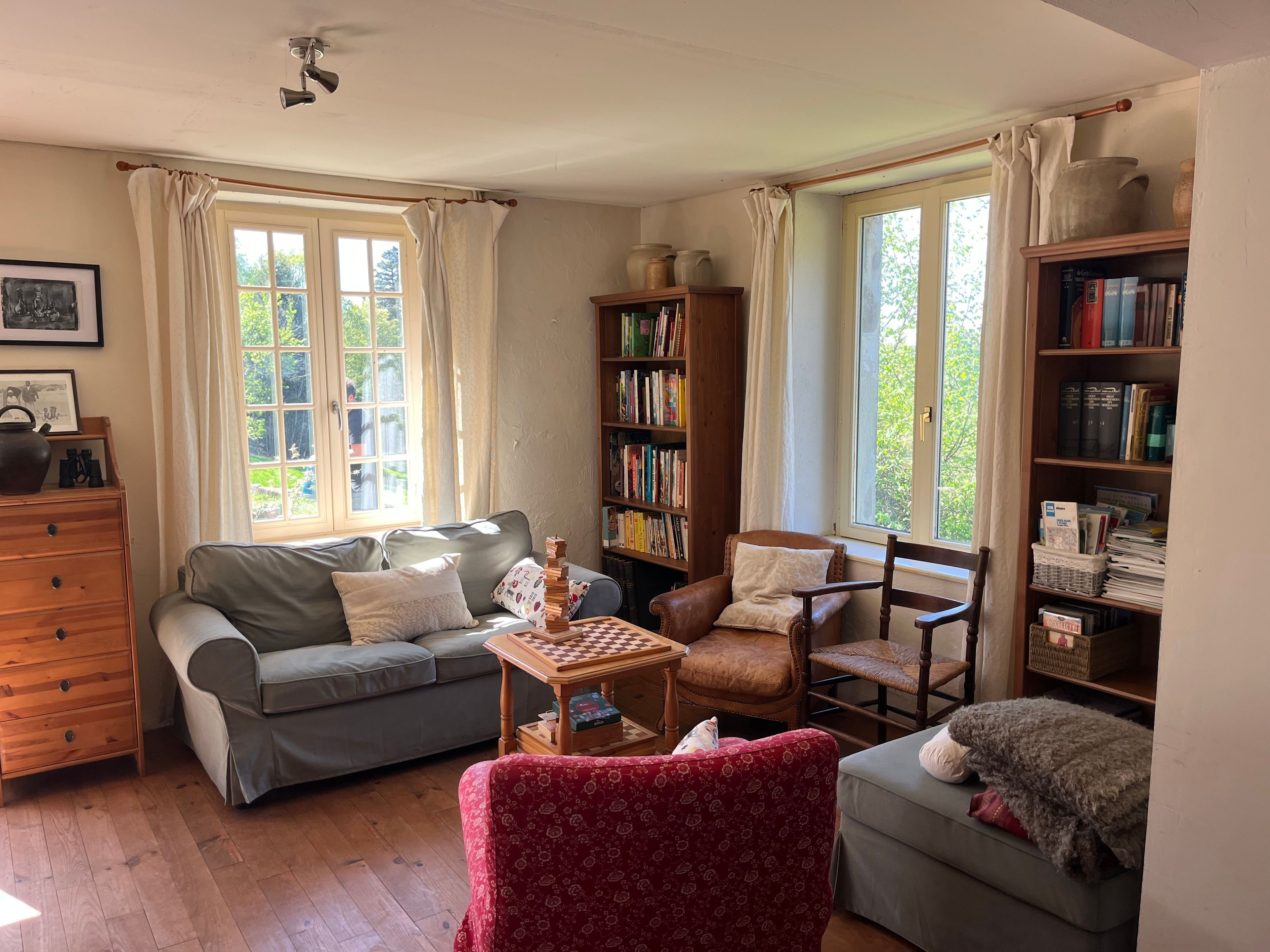 Een gezellige woonkamer met een bank, fauteuil en een kleine tafel met een schaakbord. Er staan boekenkasten vol boeken en een raam met gordijnen.