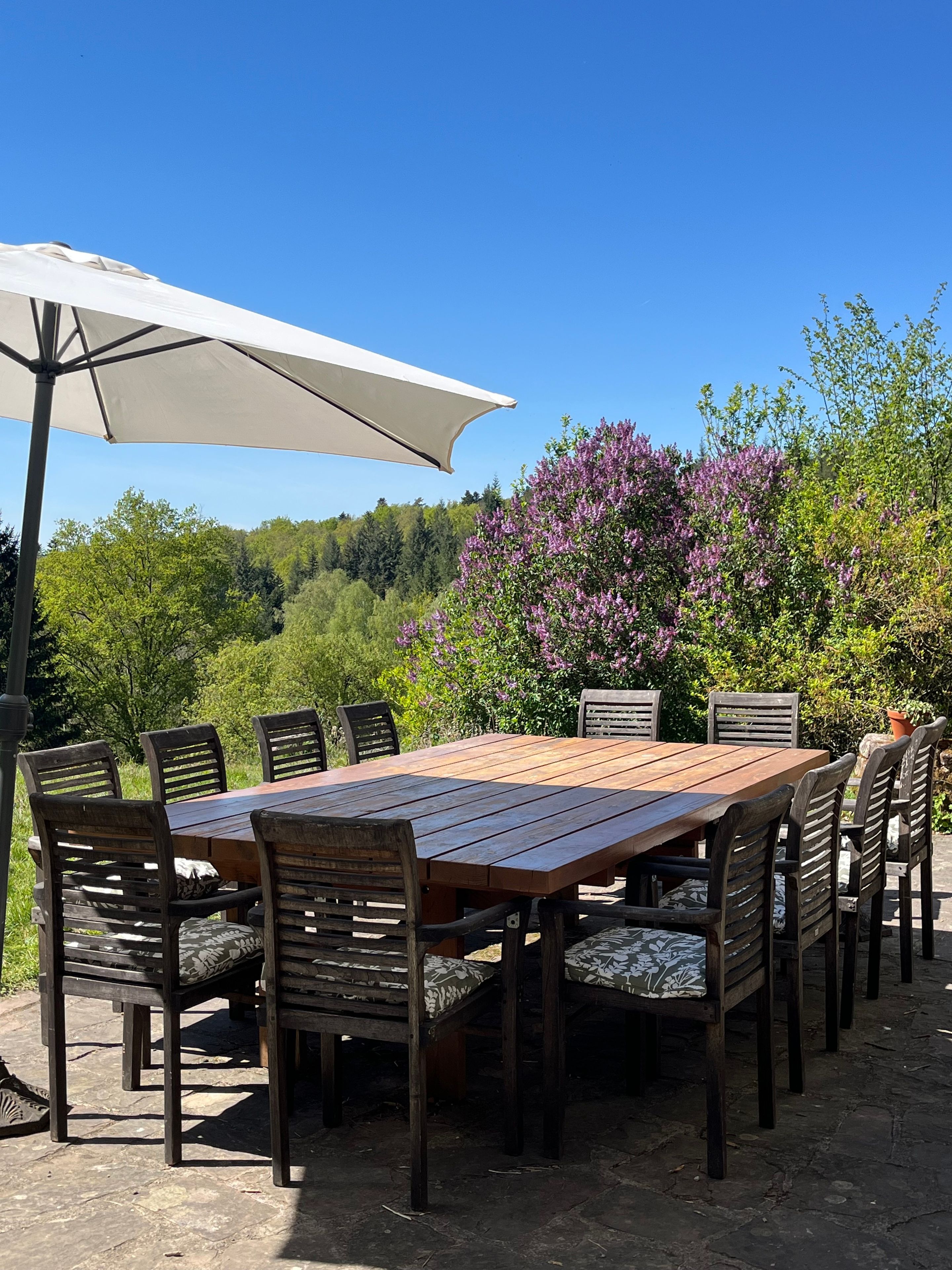 Ein Essbereich im Freien mit einem Holztisch und Stühlen, einem weißen Sonnenschirm und einer Kulisse aus Bäumen und violetten Blumen.