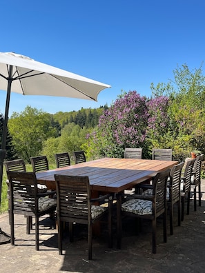 Ein Essbereich im Freien mit einem Holztisch und Stühlen, einem weißen Sonnenschirm und einer Kulisse aus Bäumen und violetten Blumen.