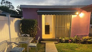 Um imóvel residencial com um alpendre, mesa de vidro, cadeiras, um pequeno jardim com plantas em vasos e uma porta que dá para o interior.