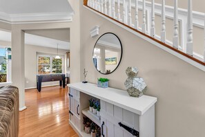 Amplia sala de estar con suelo de madera, una consola blanca con objetos decorativos, un espejo redondo y una escalera con barandilla blanca.