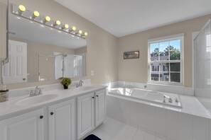 Un baño moderno con lavamanos dobles, un espejo grande con luces, una bañera y una ventana con vistas a casas.