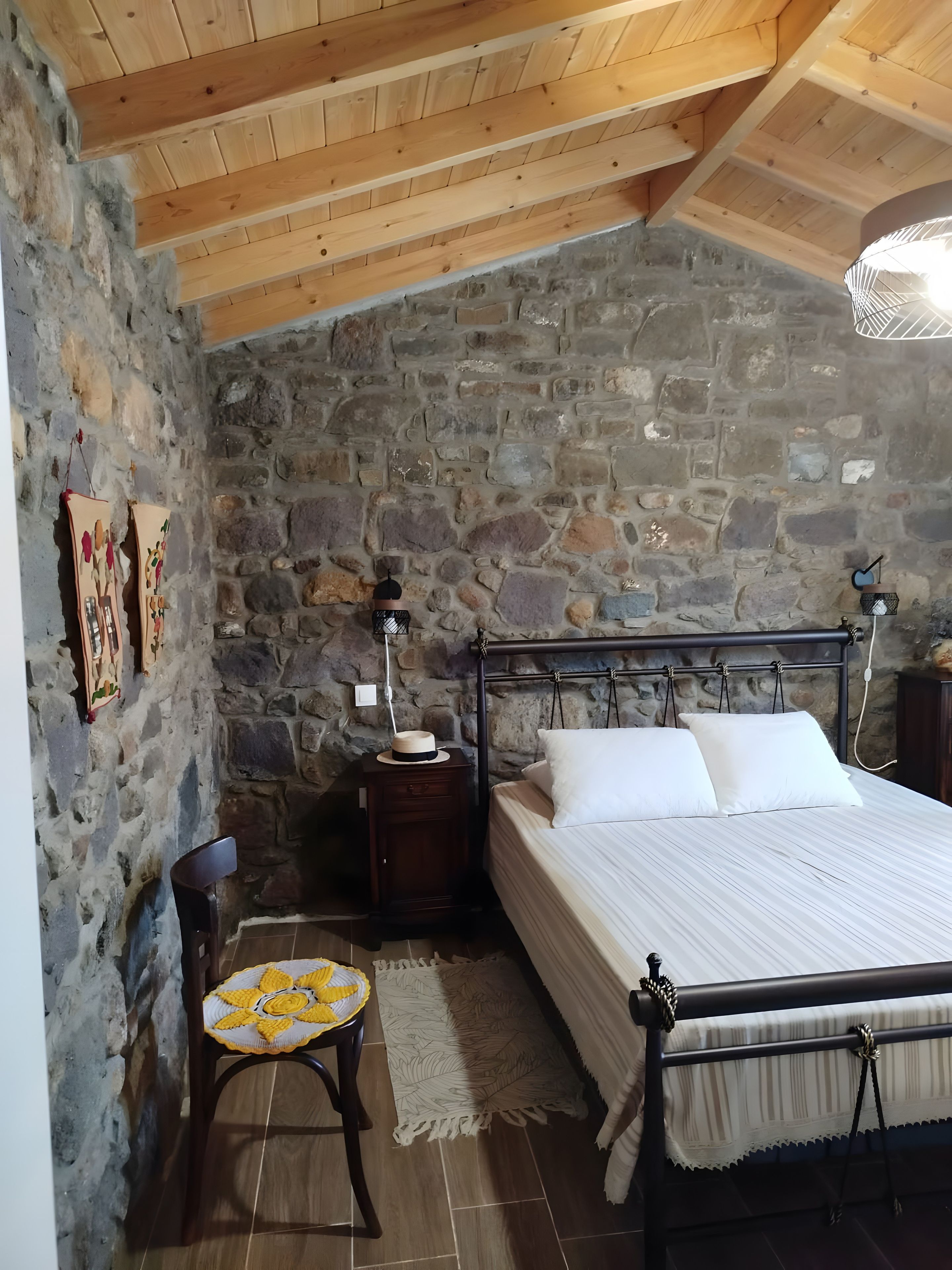 Un dormitorio con pared de piedra, vigas de madera en el techo, una cama con ropa blanca, una mesita de noche, una silla con cojín amarillo y una lámpara colgante.