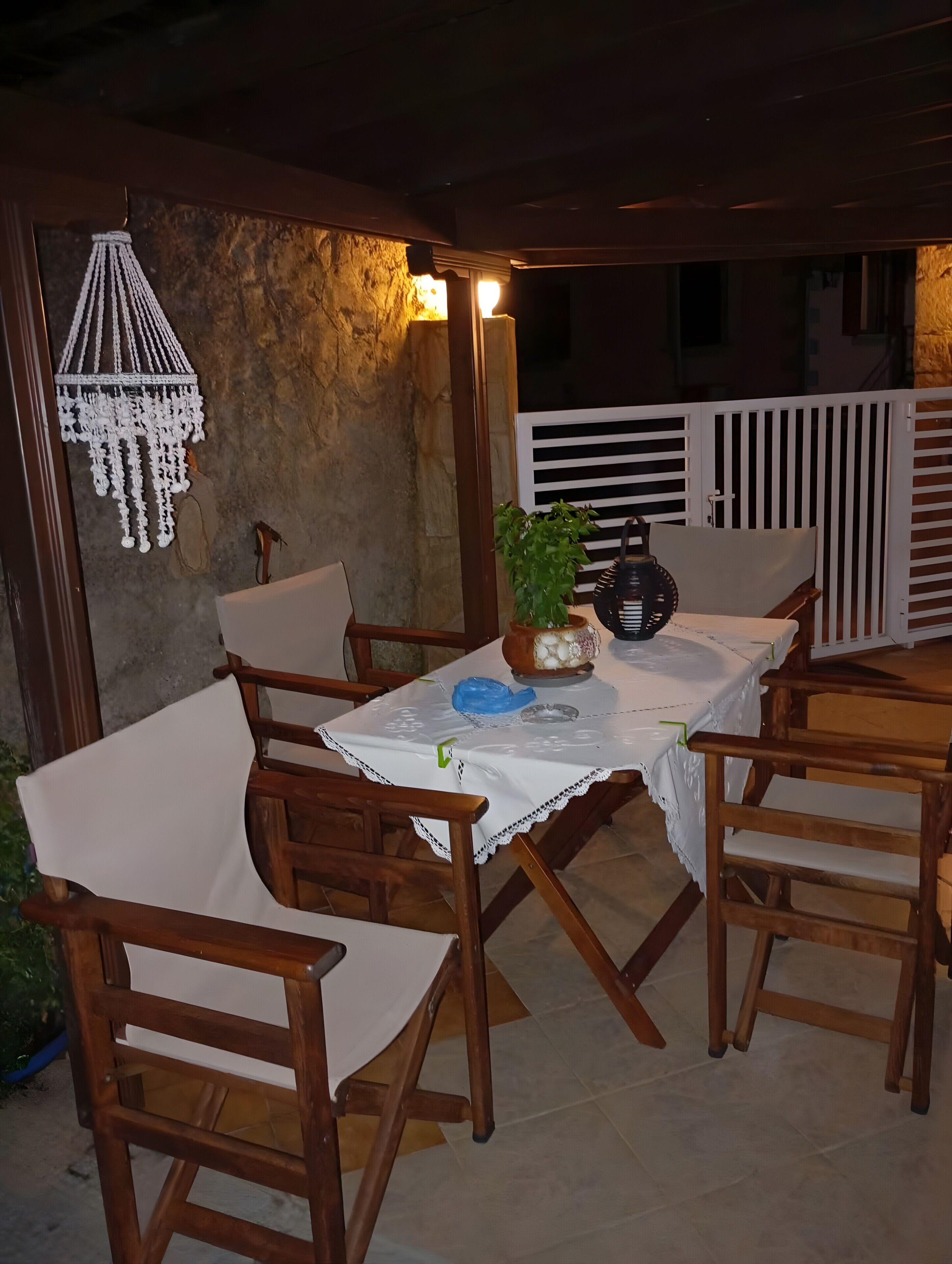 Un área de comedor al aire libre con una mesa dispuesta para cuatro, rodeada de sillas de madera, una planta en maceta y una lámpara colgante decorativa.