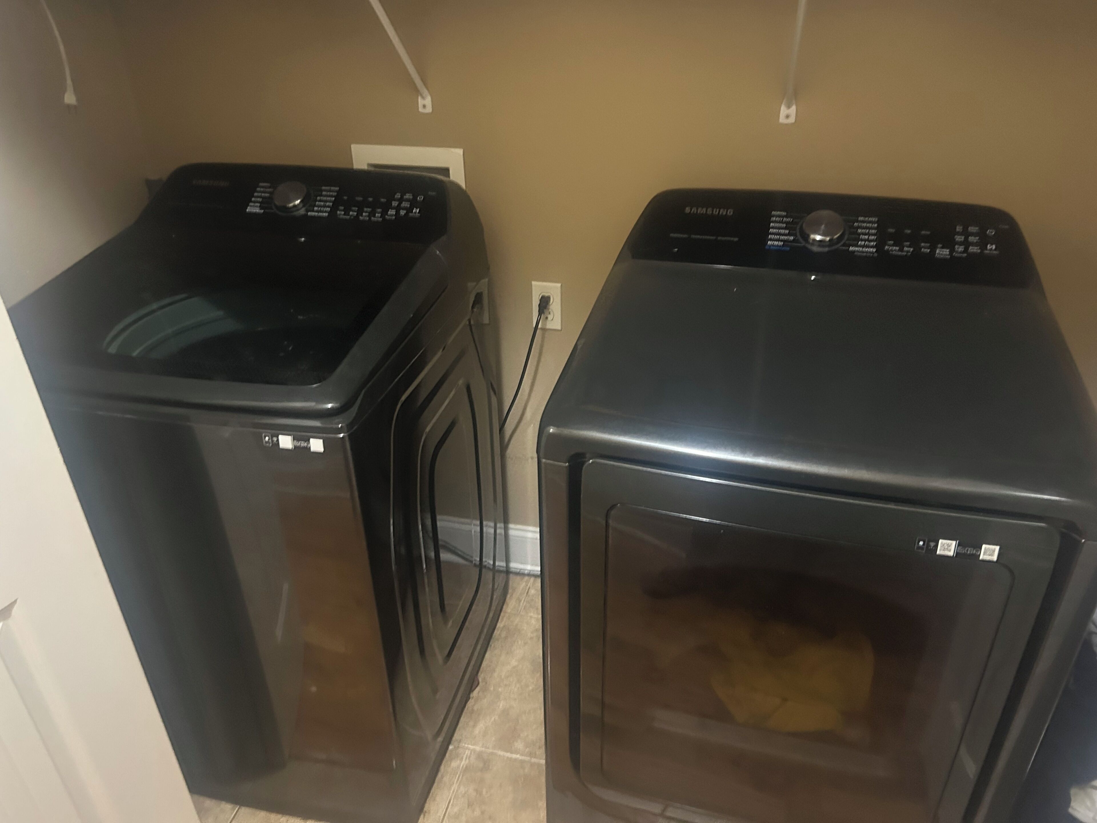 On aperçoit deux appareils électroménagers noirs, probablement un lave-linge et un sèche-linge, dans une pièce aux murs de couleur beige et au sol carrelé.