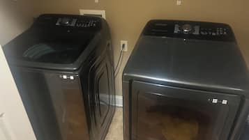 On aperçoit deux appareils électroménagers noirs, probablement un lave-linge et un sèche-linge, dans une pièce aux murs de couleur beige et au sol carrelé.