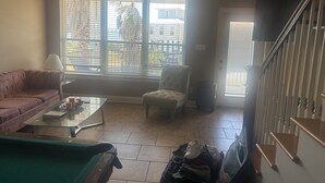 Ein Wohnzimmer mit einer Couch, einem Sessel und einem Glastisch. Es gibt ein Fenster mit Jalousien, einen Deckenventilator und eine Treppe mit Holelllauf.