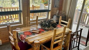 Une table à manger en bois, entourée de six chaises, un vase rempli de fleurs et une vue sur l’extérieur grâce à de grandes fenêtres.