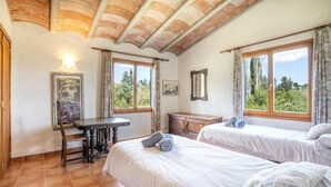 A room with two beds, a wooden desk, and a window with a view of trees.