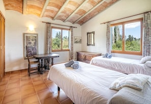 A room with two beds, a wooden desk, and a window with a view of trees.