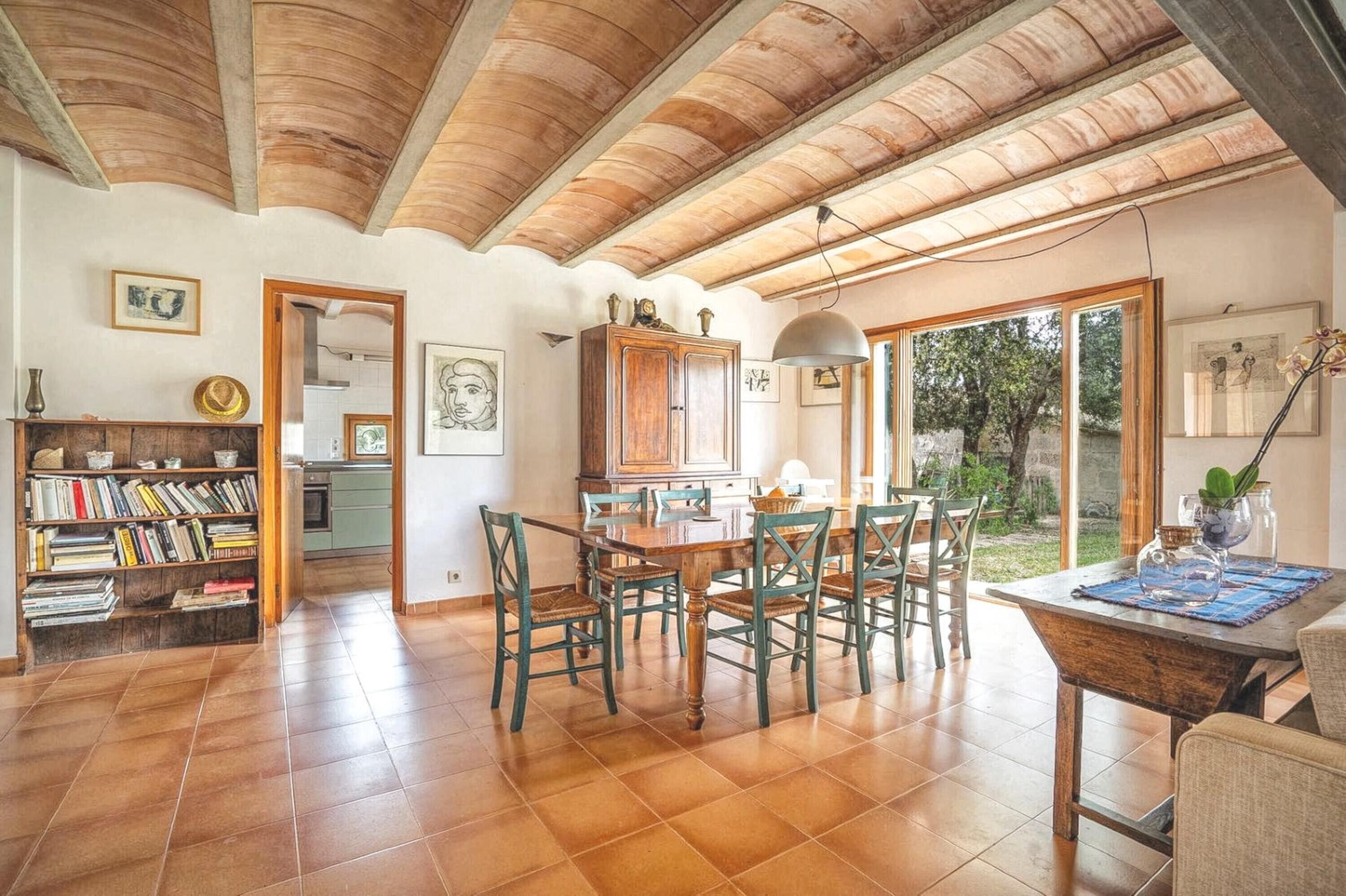 A spacious dining area with a wooden table and chairs, a kitchen in the background, and a view of the outdoors through large windows.