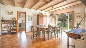 A spacious dining area with a wooden table and chairs, a kitchen in the background, and a view of the outdoors through large windows.