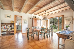 A spacious dining area with a wooden table and chairs, a kitchen in the background, and a view of the outdoors through large windows.
